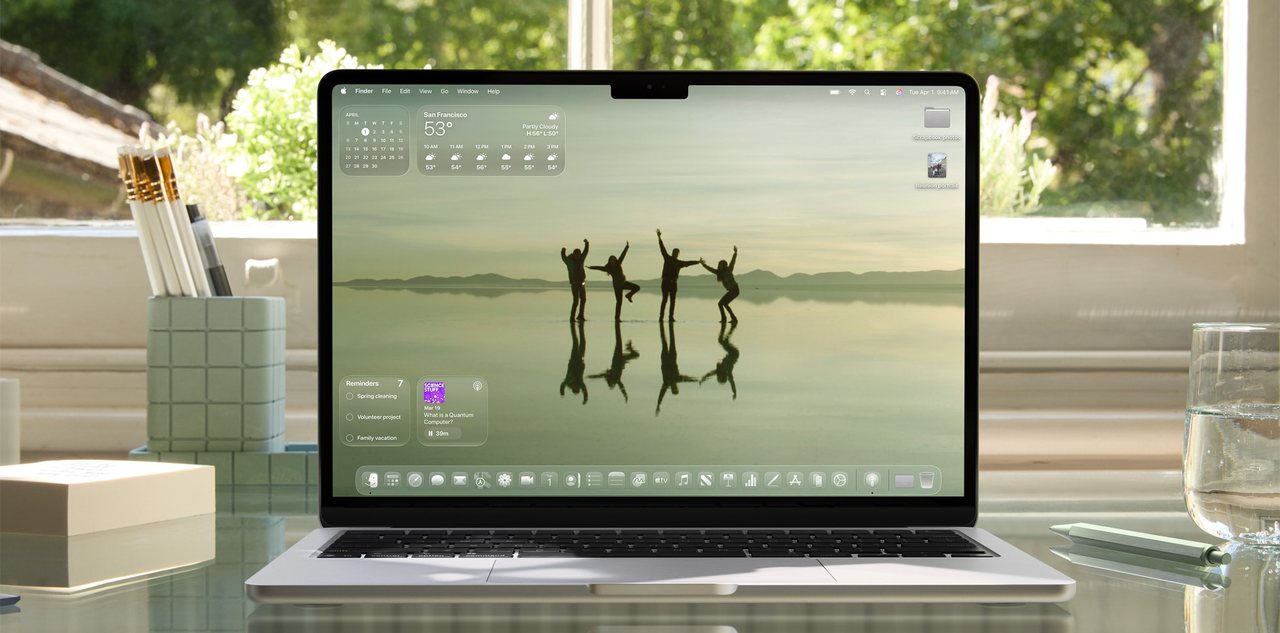 A MacBook on a desk with the translucent Liquid Glass interface on its screen